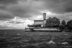 Schwarz Weiß Foto von Schloss Montfort in Langenargen am Bodensee. Das Schloss ist von Bäumen umgeben, auf dem Turm weht eine Flagge. Im Vordergrund sind vom Wind aufgepeitschte Wellen, am Himmel sind dunkle Wolken.